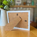 White picture frame with a kickstand on brown table next to a jug of flowers